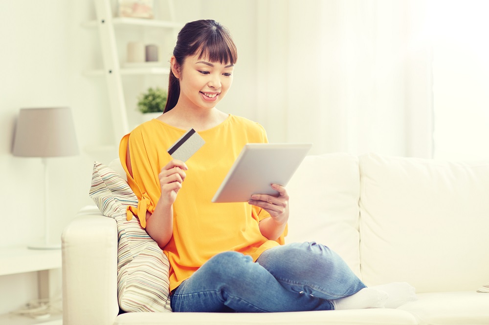 people, internet bank, online shopping, technology and e-money concept - happy asian young woman sitting on sofa with tablet pc computer and credit card at home